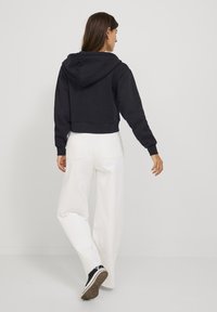Hoodie preto cortado com capuz ajustável, combinado com calças brancas largas e sapatilhas pretas, visto por trás.