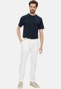 T-shirt in cotone navy, pantaloni bianchi, cintura nera con accenti metallici, sneakers beige. Modello in piedi con una mano in tasca, sfondo neutro.