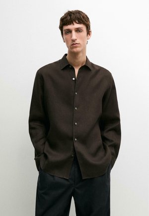 Giovane uomo con capelli castani corti indossa una camicia marrone scuro con bottoni e pantaloni neri, in piedi con le mani nelle tasche contro uno sfondo chiaro.