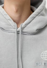 Felpa leggermente grigia con cappuccio e lacci, caratterizzata da una texture morbida e un design del logo frontale. Il colletto è rotondo con un leggero bordo a coste.