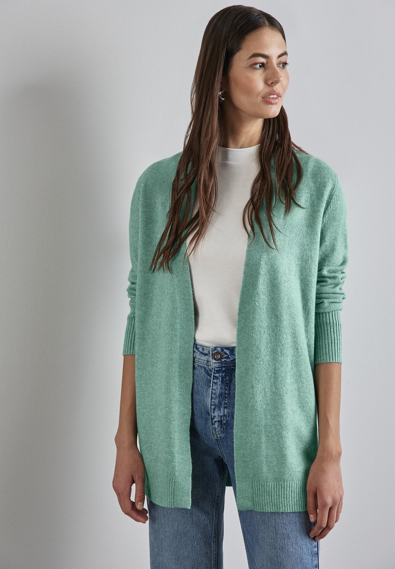 Hellgrüner Cardigan aus weichem Material, mit geripptem Saum und Bündchen, offenem Design, getragen über einem cremefarbenen Oberteil und blauen Jeans.