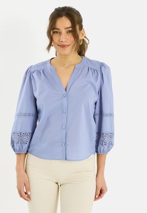 Mujer con blusa azul clara abotonada con mangas abullonadas y detalles de encaje, combinada con pantalones beige, posando frente a un fondo liso.