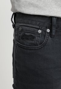 Vaqueros de mezclilla negra con un acabado texturizado, que cuentan con un diseño de cinco bolsillos, botones plateados y un logo bordado en el bolsillo delantero.