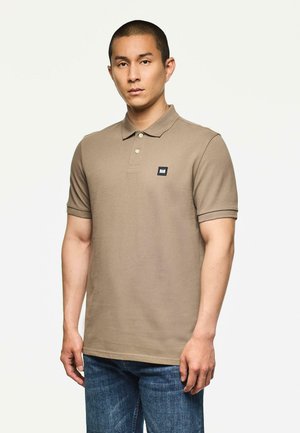 Man staat gekleed in een taupe polo met korte mouwen, voorzien van twee knopen en een klein zwart logo op de patch, gecombineerd met een paar blauwe jeans.