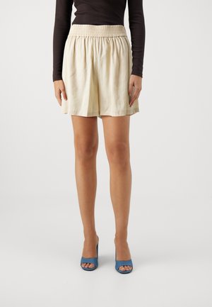 Cremefarvede shorts med en samlet elastisk talje, der har en løs pasform og blødt, let stof, kombineret med blå hælede mules.