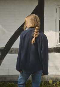 Laivansinisestä villasekoitekankaasta valmistettu takki, jossa on rento leikkaus, suuri kaulus ja roikkuvat olkapäät. Yhdistetty vaaleansinisiin farkkuihin. Luonnollinen tausta.
