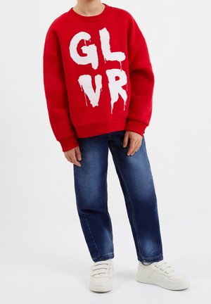 Sweatshirt rouge avec le texte "GLVR" en style graffiti blanc, associé à un jean en denim bleu et des baskets blanches à trois bandes Velcro.
