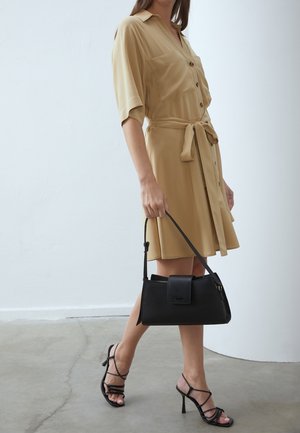 Beige blousejurk met korte mouwen en een ceintuur in de taille, gecombineerd met een zwarte handtas en zwarte hooggehakte sandalen met bandjes.