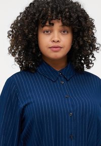 Camicia blu navy con bottoni, presenta strisce verticali bianche, un colletto classico e un tessuto liscio e testurizzato.