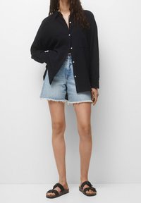 Camicia nera con bottoni, dal taglio rilassato, maniche lunghe e spacchi laterali, abbinata a pantaloni denim e sandali neri.