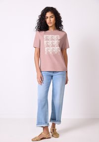 Rosa bomull t-shirt med ett fyrkantigt dinosauriemönster, tillsammans med ljusblå vida jeans och gula sandaler med utskärningar.