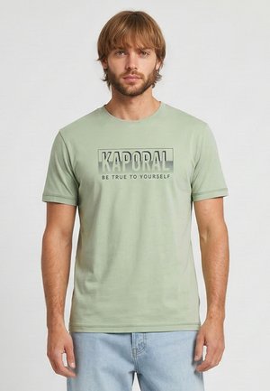 Homme aux cheveux châtain clair et à la barbe, portant un t-shirt Kaporal vert sauge avec le texte « Sois fidèle à toi-même », assorti à un jean bleu clair.