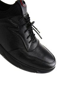 Zapatilla de cuero negra con parte superior texturizada, cordones redondeados, forma elegante y un acento en contraste en el ojal. Suela de goma con un patrón de tracción distintivo.