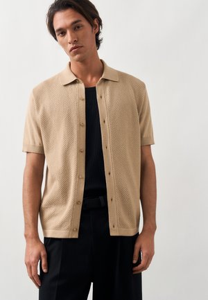 Man die een beige kort mouws vest met knopen draagt over een zwart geribd shirt en zwarte broek, staand tegen een effen witte achtergrond.
