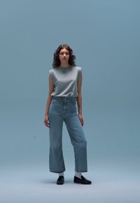 Top cinzento sem mangas combinado com jeans de perna larga azul claro de cintura alta, complementado por sapatos escuros e meias brancas contra um fundo azul simples.