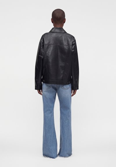 Personne de dos, portant une veste en cuir noire et un jean bleu clair évasé à ourlets effilochés, sur fond uni clair.