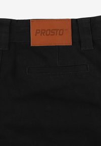 Czarne materiałowe spodnie o gładkiej fakturze, z brązową skórzaną naszywką z napisem "PROSTO" oraz jedną tylną kieszenią.