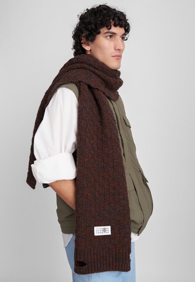 Cachecol de malha castanho com um padrão texturizado, apresentando acentos escuros e um rótulo tecido, pendurado sobre uma pessoa vestindo um colete verde-oliva.
