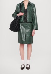 Zelená bunda z umělé kůže se širokými rukávy, doplněná odpovídající sukní s předním rozparkem. Černá pletená taška přes rameno. Bílé ponožky a pantofle.