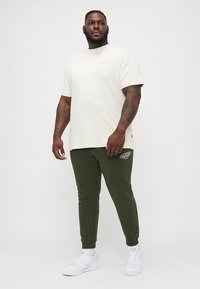 Kremfarget t-skjorte med korte ermer og subtilt trykk, kombinert med mørkegrønne joggebukser med grafisk aksent, fullført med hvite joggesko.