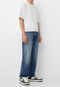 Valkoinen lyhythihainen t-paita yhdistettynä vaaleansinisiiin, väljälahkeisiin farkkuihin ja mustavalkoisiin matalakärkisiin tennareihin, joissa on teksturoitu kuvio.