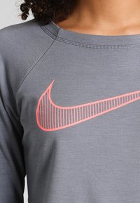 Maglietta sportiva grigia a maniche lunghe con scollo rotondo; presenta un logo Nike rosa ben visibile con strisce orizzontali. Tessuto morbido e liscio.