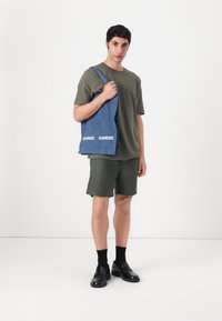 Denim-Tote-Bag mit weißem "SAMSØE"-Logo, gehalten von einer Person in einem grünen T-Shirt und Shorts, vor einem schlichten weißen Hintergrund stehend.