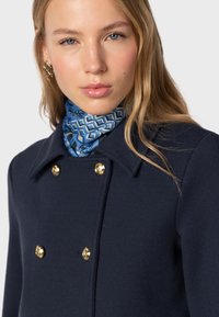 Navy coat with gold buttons, featuring a structured collar. The model wears a blue patterned scarf and has wavy, light brown hair.