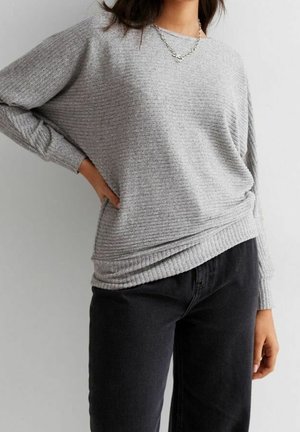 Personne portant un pull tricoté côtelé gris clair à manches longues et un pantalon noir, avec une main posée sur la hanche.