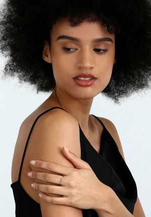 Schwarzes Satin-Oberteil mit drapierter Gestaltung. Nahaufnahme von Händen, die einen silbernen Ring mit einem zentralen Stein präsentieren. Die Modellent hat lockiges schwarzes Haar und ein neutrales Make-up.