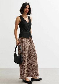 Schwarzes, strukturiertes ärmelloses Oberteil mit Knöpfen, kombiniert mit weit geschnittenen, beige-braun getupften Leopardenhosen; eine schwarze Handtasche und Ballerinas runden das Outfit ab.