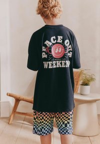 Svart oversized t-shirt med grafisk text och ett smiley-motiv på baksidan. Färgglada rutiga shorts i gradientpastellnyanser. Inomhusmiljö.