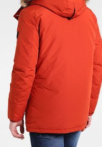 Červená zateplená bunda s kapucí lemovanou kožešinou, viditelným prošitím a elastickými manžetami. Nošena s modrými džínami, které zdůrazňují uvolněný střih.