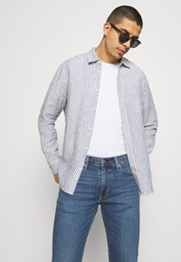 Camisa ligera, a rayas azul y blanca, de diseño abotonado, con mangas largas, llevada sobre una camiseta blanca, combinada con unos vaqueros de mezclilla azules.