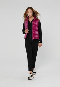 Magenta puffer vest met een glanzende afwerking, ritssluiting en zijzakken, gedragen over een zwart top en zwarte broek, gecombineerd met zwart-gouden sneakers.