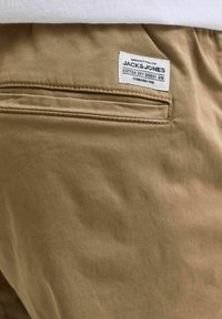 Pantalons en coton beige avec une poche arrière. Présente une étiquette avec le texte "JACK & JONES". Texture lisse avec un léger éclat.