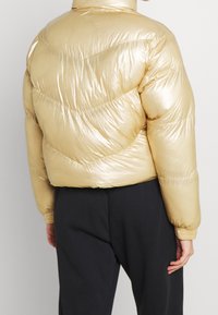 Złota kurtka puchowa o lśniącej, gładkiej teksturze, z ukośnym przeszyciem i elastycznymi mankietami. Krótki krój, lekki materiał.
