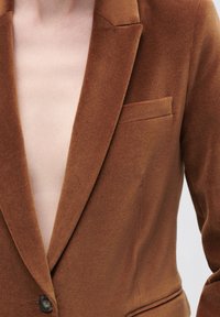 Brauner Samtblazer mit tiefem V-Ausschnitt, Reverskragen, einem Knopfverschluss und einer Brusttasche. Weiche Textur und taillierte Passform.