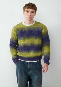 Gebreide trui in levendig groen en paarse horizontale strepen, met een ribbelstructuur en een ronde hals. Draagt over een wit shirt en gecombineerd met denim.