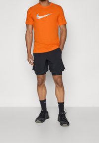 Camiseta Nike naranja con un logo azul y rosa, combinada con pantalones cortos deportivos negros y zapatillas negras, con detalles texturizados.