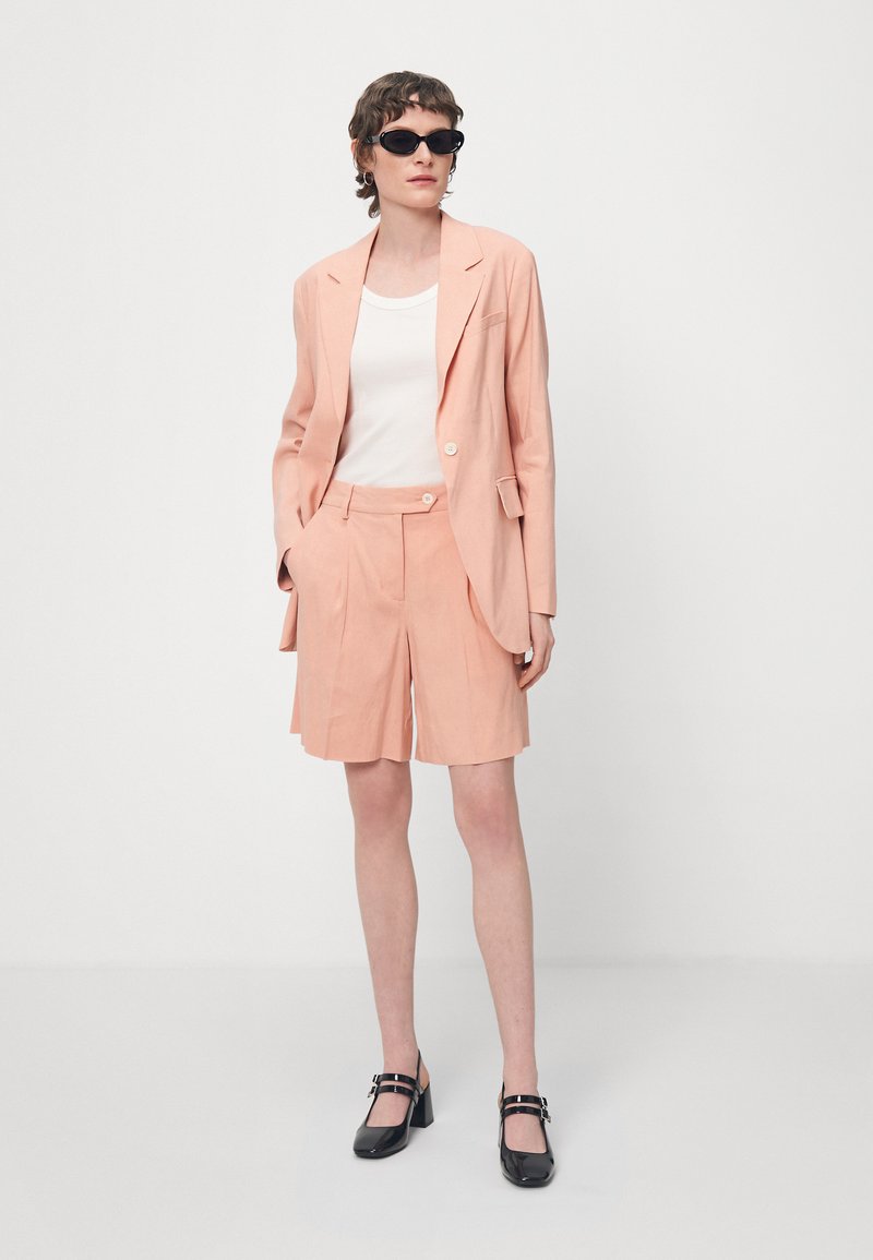 Pfirsichfarbener Blazer mit passenden Shorts, leichter Stoff, taillierte Passform, weißes Oberteil darunter und schwarze Riemchenheels. Accessoires mit Sonnenbrille.