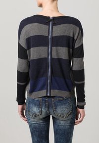 Randig stickad tröja i grått och marinblått med en dragkedja bak, med ett ribbat nederkant och ärmslut. Kombinerad med bleka blå jeans.