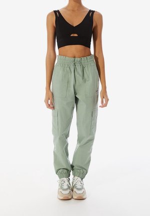 Woman wearing black cropped tank top, light green cargo pants with elastic cuffs, and beige-white sneakers, standing straight on white background.