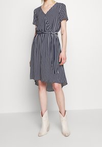 Navy and white striped dress with a V-neck, short sleeves, and a belted waist; knee-length with a longer back hem. Worn with white ankle boots.