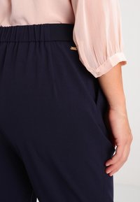 Marinblå, elastiska byxor med resårmidja och sidofickor, kombinerat med en persikofärgad, veckad blus med knäppte ärmslut.
