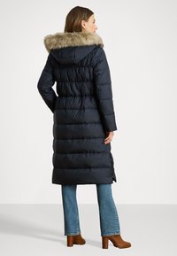 Sötétkék puffermellény műszőrme kapucnival, hosszú sziluettel, steppelt textúrával és oldalsó hasítékokkal, világoskék farmernadrág és barna magassarkú csizma felett viselve.