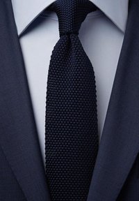 Marinblå stickad slips, texturerat tyg, med ett enhetligt mönster. Bärs över en ljusblå skjorta, i kombination med en mörk kavaj, rena linjer.