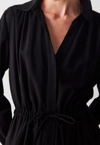 Nainen, jolla on päällä musta pitkähihaisesta puserosta, jossa on kaulus ja nyöri vyötäröllä.