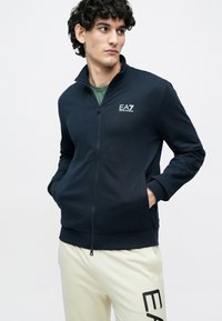 Tmavomodrá bunda na zips z mäkkej tkaniny s vysokým golierom, bočnými vreckami a malým logom EA7 na hrudi. Klasický strih.