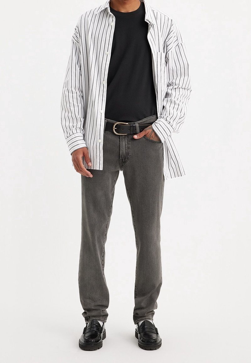Homme portant un t-shirt noir, une chemise boutonnée rayée blanche, un jean gris avec ceinture noire, et des mocassins noirs, debout avec une main dans la poche.